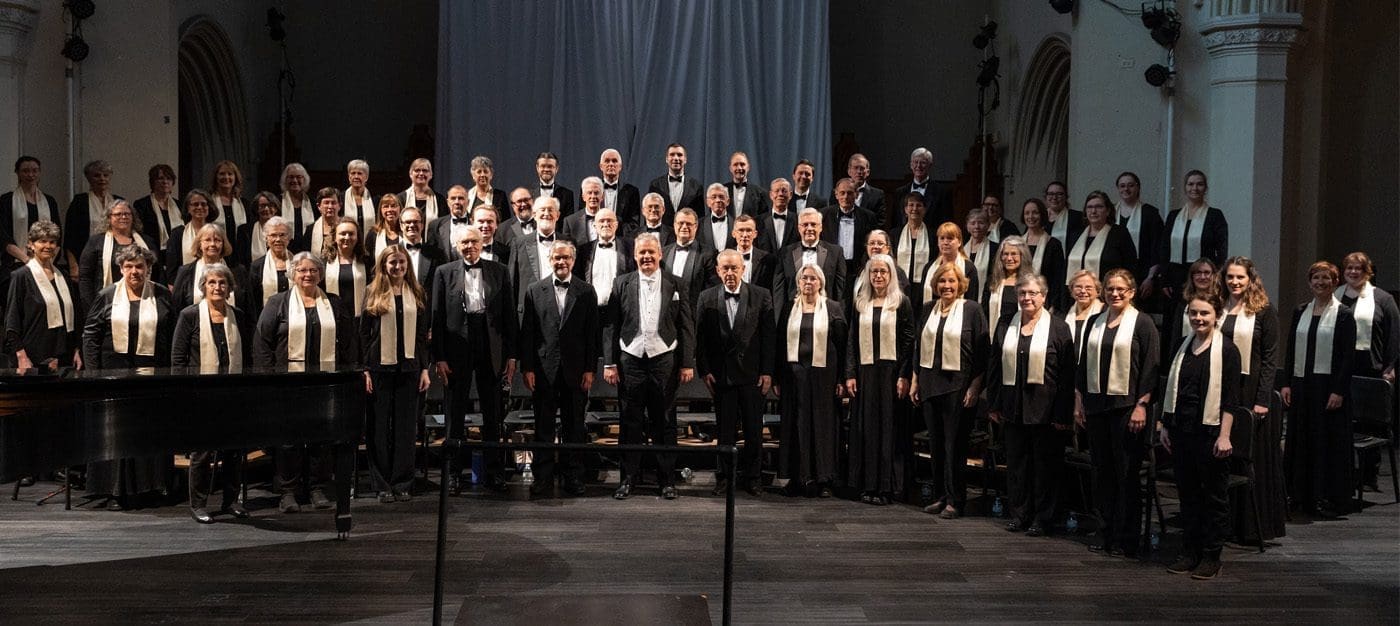 group1 A large choir in black-and-white formal attire performs on a stage with a pianist at the left and a conductor at the center.