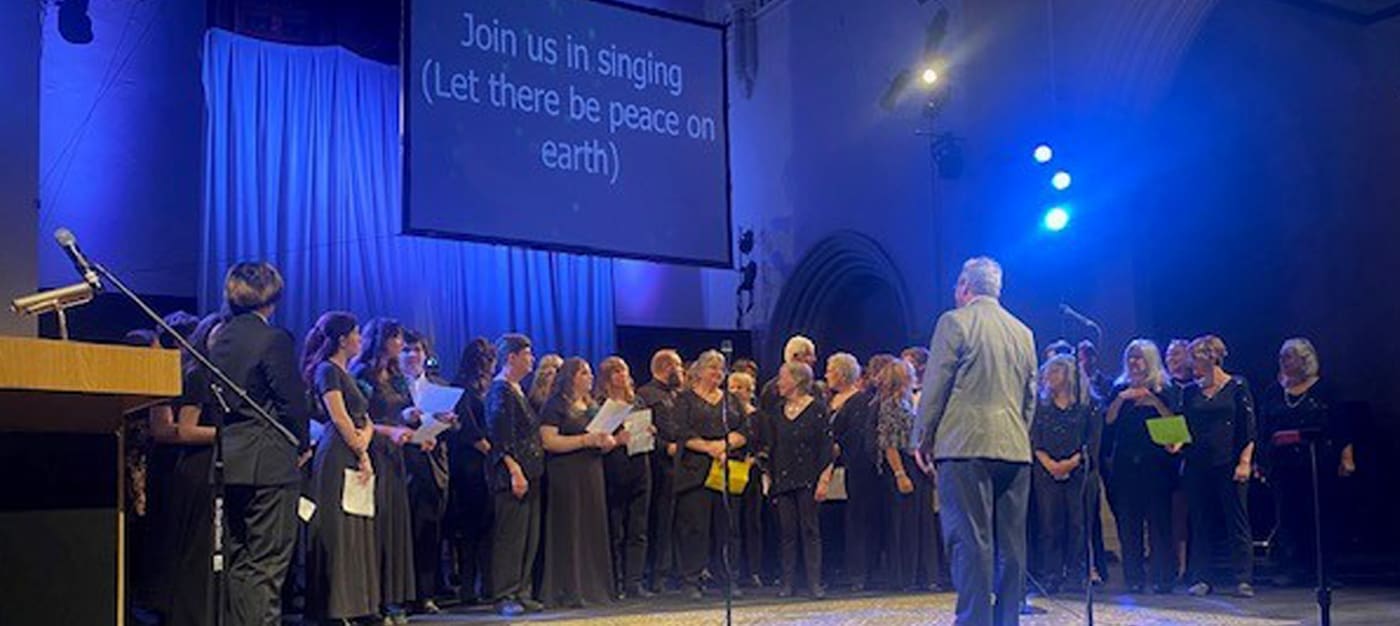 bbotb-3 A choir performs on a stage under blue lighting, led by a conductor, with a screen displaying lyrics in the background.