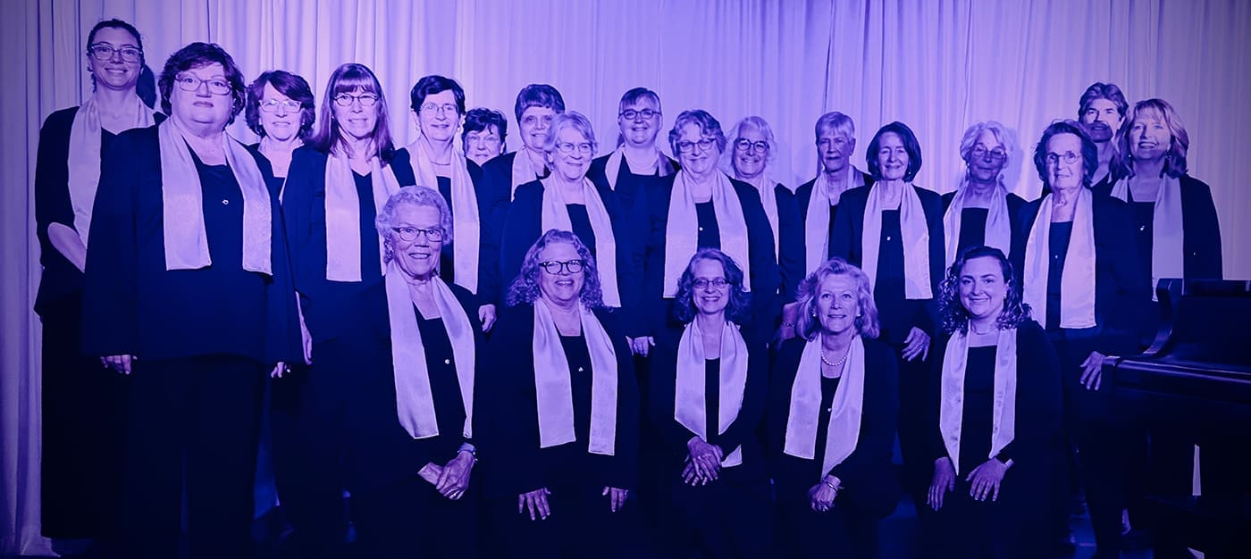 altos2 A large choir of men and women in black outfits with light scarves posing on a stage, with a piano visible on the right.