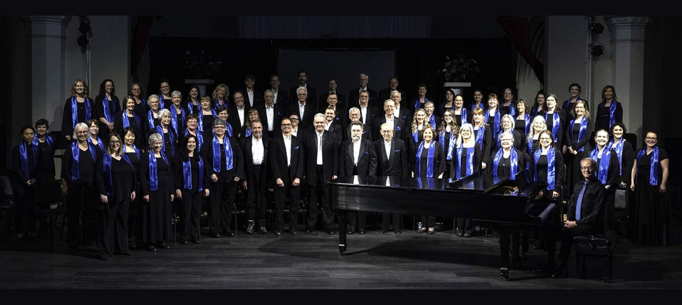 may-2025 A large choir in black attire with blue accents performs on a stage beside a grand piano, guided by a conductor.